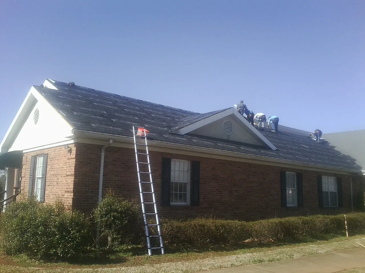 Crew tearing shingles off a brick ranch roof with a ladder set — Metal Roof Repair work by our crew in Moneta