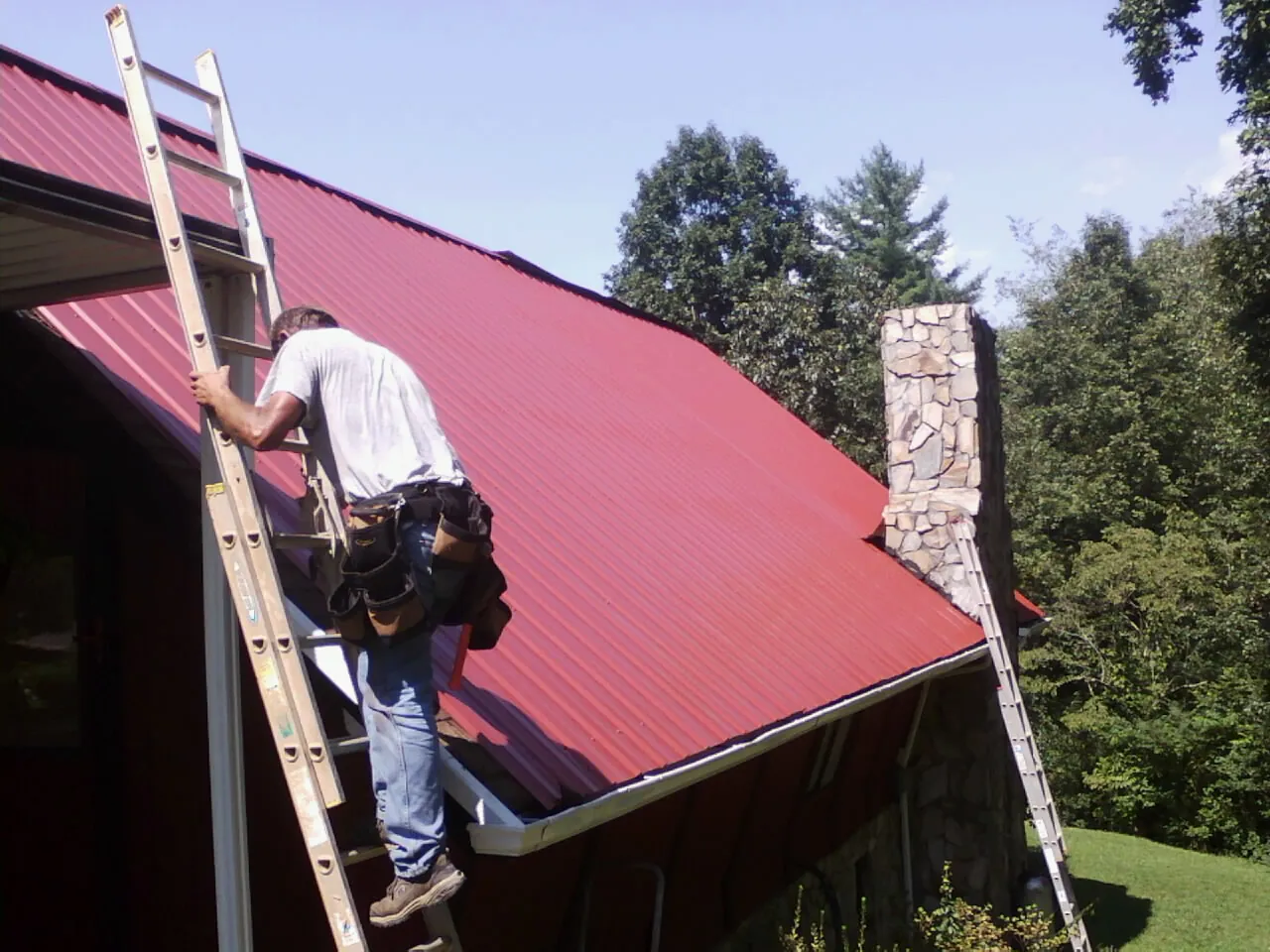 Roofer on a ladder beside a red metal roof and stone chimney — Soffit & Fascia Repair by Roofing Brothers in Moneta